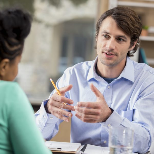 Confident mid adult Caucasian manager interviews prospective African American female employee. The man is gesturing while talking. The woman's back is to the camera.